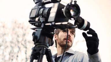 Ein Kameramann richtet eine professionelle Videokamera aus und überprüft die Einstellungen. Die Szene zeigt eine eigenständige Tätigkeit in der Medienproduktion.