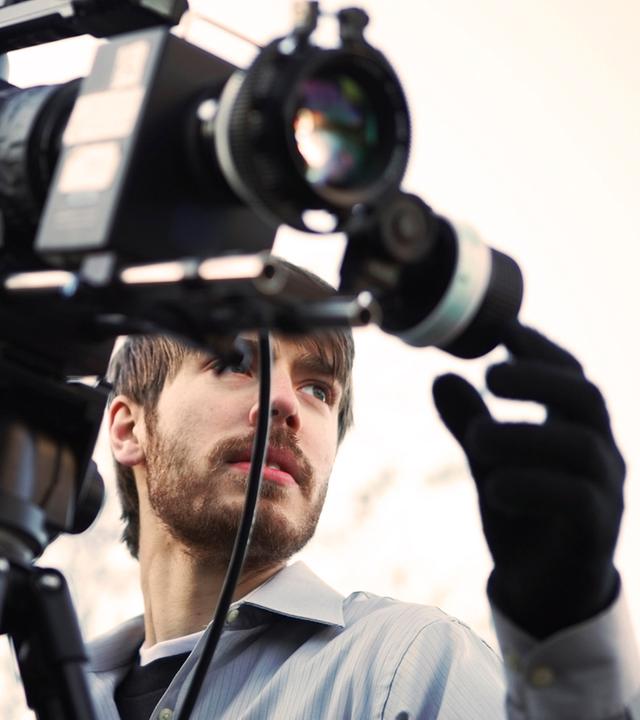 Ein Kameramann richtet eine professionelle Videokamera aus und überprüft die Einstellungen. Die Szene zeigt eine eigenständige Tätigkeit in der Medienproduktion.