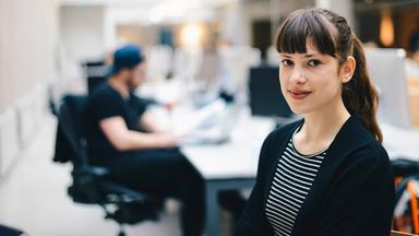 Eine Frau steht im Vordergrund eines modernen Büros und blickt in die Kamera, während im Hintergrund eine weitere Person am Computer arbeitet. Die Szene vermittelt einen Einblick in den Arbeitsalltag im Team.