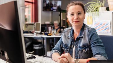 Eine Frau sitzt an einem Arbeitsplatz im Büro und blickt aufmerksam in Richtung Kamera. Im Hintergrund sind weitere Arbeitsplätze zu sehen, die eine redaktionelle Umgebung andeuten.