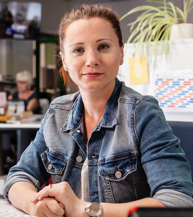 Eine Frau sitzt an einem Arbeitsplatz im Büro und blickt aufmerksam in Richtung Kamera. Im Hintergrund sind weitere Arbeitsplätze zu sehen, die eine redaktionelle Umgebung andeuten.