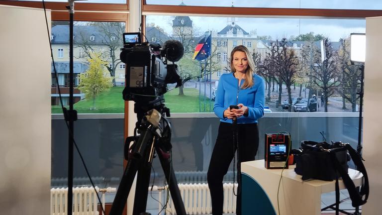 Sarah Take mit Handy in der Hand in einem Fernsehstudio, im Hintergrund sieht man aus dem Fenster das Bundesverfassungsgericht