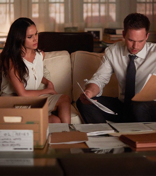"Suits - Karten auf den Tisch": Rachel (Meghan Markle) und Mike (Patrick J. Adams) sitzen nebeneinander auf einem Sofa und schauen Dokumente an, die Mike in beiden Händen hält. Beide haben einen besorgten Gesichtsausdruck. Vor ihnen auf dem Tisch eine offene Kiste und ausgebreitete Dokumente.