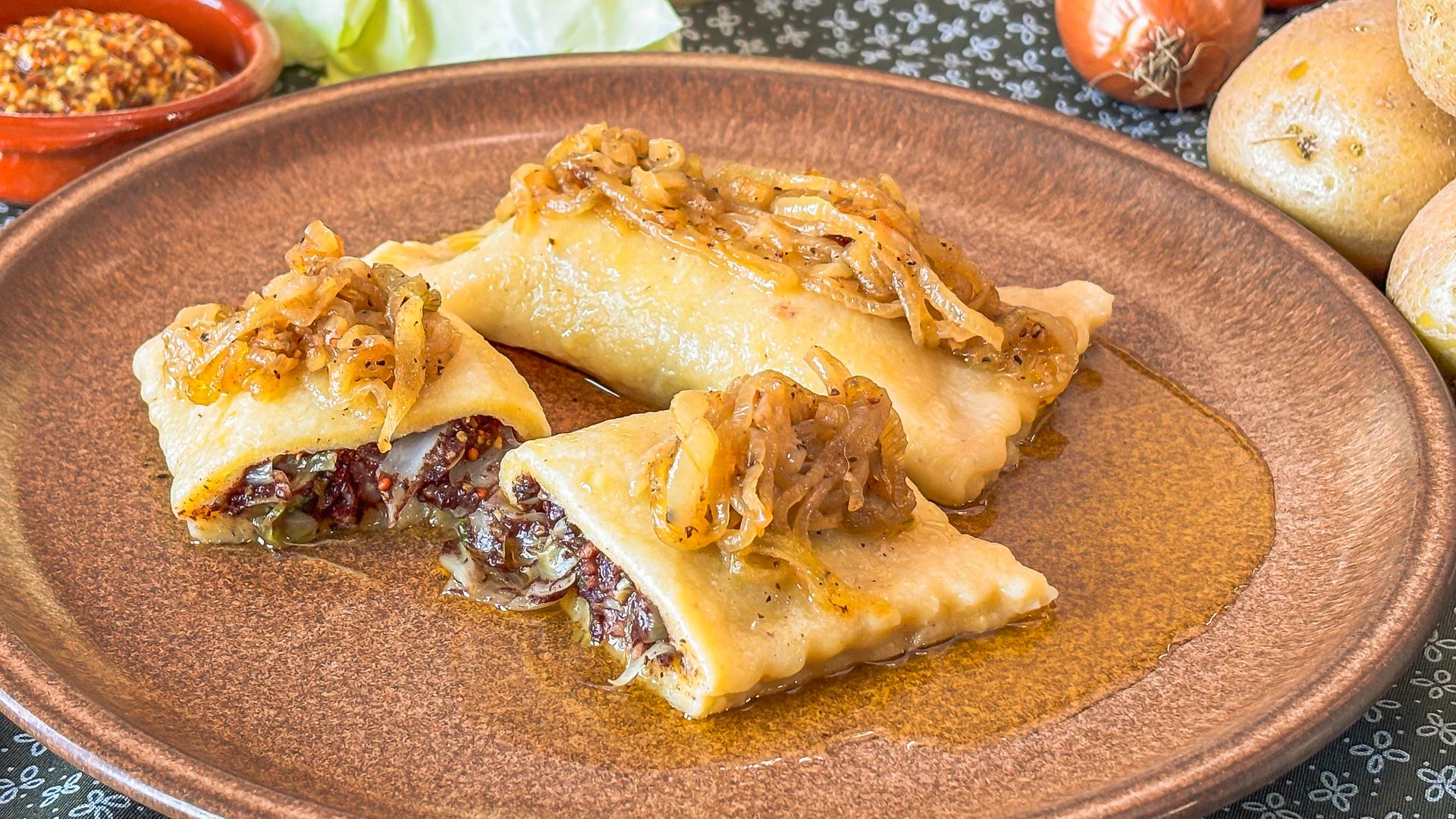 Brauner Teller mit Kartoffelmaultaschen, gefüllt mit Blutwurst und getoppt mit Schmorzwiebeln.