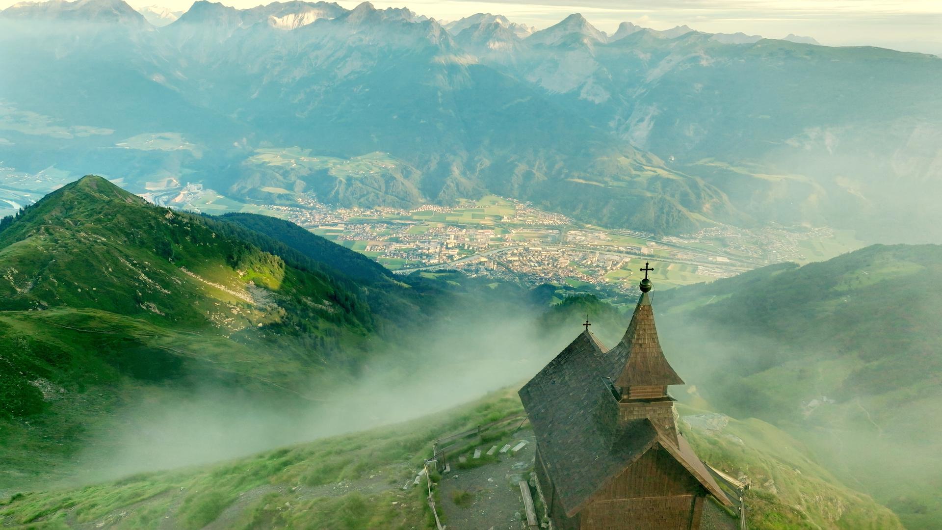 Das Bild zeigt die Kellerjochkapelle, die sich auf einem Bergvorsprung im Karwendelgebirge befindet. Die Kapelle hat einen traditionellen, schindelgedeckten Dachstuhl und ist mit einem kleinen Kreuz auf der Spitze versehen. Vor der Kapelle erstreckt sich eine grüne, hügelige Landschaft, die sanft abfällt und in die darunter liegende Stadt Schwaz übergeht. Im Hintergrund sieht man die majestätischen Gipfel der umliegenden Berge, die teilweise in Nebel gehüllt sind. Die Landschaft ist von einer dichten Vegetation geprägt, die zwischen Wald- und Wiesengebieten wechselt. Am Fuß des Berges sind die Strukturen der Stadt Schwaz und landwirtschaftliche Flächen zu erkennen, die sich in der Talsohle ausbreiten. Die gesamte Szene vermittelt ein Gefühl von Ruhe und Erhabenheit, während die Wolken und der Nebel eine mystische Atmosphäre schaffen.