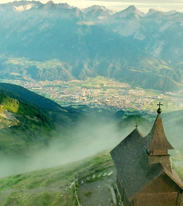 Das Bild zeigt die Kellerjochkapelle, die sich auf einem Bergvorsprung im Karwendelgebirge befindet. Die Kapelle hat einen traditionellen, schindelgedeckten Dachstuhl und ist mit einem kleinen Kreuz auf der Spitze versehen. Vor der Kapelle erstreckt sich eine grüne, hügelige Landschaft, die sanft abfällt und in die darunter liegende Stadt Schwaz übergeht. Im Hintergrund sieht man die majestätischen Gipfel der umliegenden Berge, die teilweise in Nebel gehüllt sind. Die Landschaft ist von einer dichten Vegetation geprägt, die zwischen Wald- und Wiesengebieten wechselt. Am Fuß des Berges sind die Strukturen der Stadt Schwaz und landwirtschaftliche Flächen zu erkennen, die sich in der Talsohle ausbreiten. Die gesamte Szene vermittelt ein Gefühl von Ruhe und Erhabenheit, während die Wolken und der Nebel eine mystische Atmosphäre schaffen.