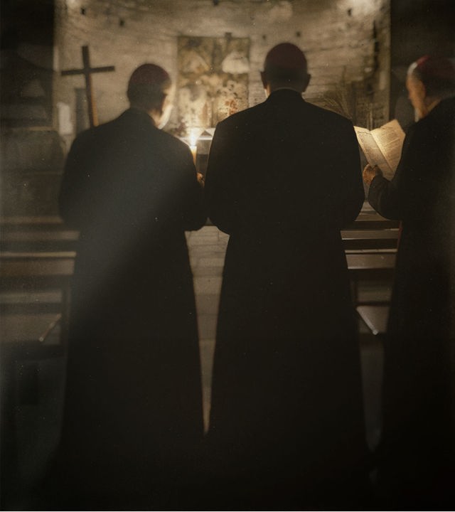 Zwei Kardinäle stehen in einer alten christlichen Katakombe und schauen auf einen Altar, über dem ein Kreuz hängt.
