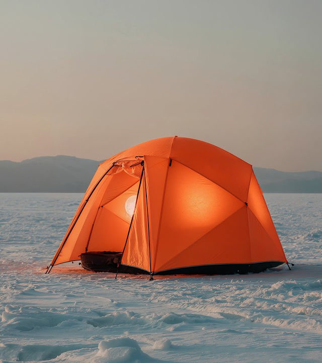 Ein orangenes Zelt steht auf einer weitläufigen verschneiten Ebene. Im Hintergrund sind Berge zu erkennen
