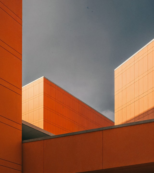 Zu sehen sind mehrere Häuserfasaden die sich wie Kuben vor dem grauen Himmel abzeichnen. Die Fasaden sind orangefarben.