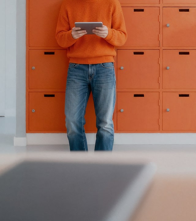 Auf dem Bild ist ein Mensch in blauer Jeans-Hose und orangenem Pullover zu sehen. Der Kopf ist außerhalb des Bildes, weshalb man sein Gesich nicht sehen kann. In seinen Händen hält er ein Tabblet. Er ist an eine Wand voll orangener Schließfächer angelehnt.