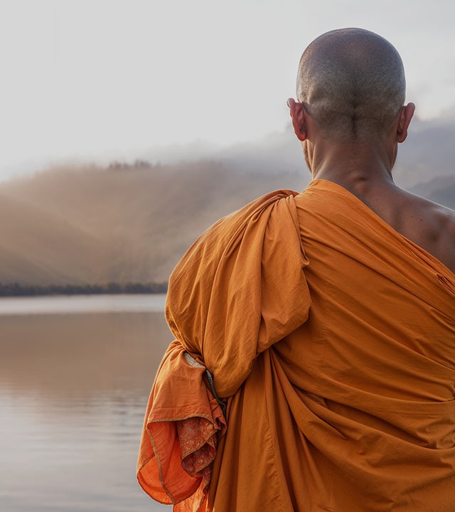 Auf dem Bild ist ein buddhistischer Mönch in orangenem Gewand zu sehen. Der Mönch ist von hinten zu sehen und schaut in die Ferne, in der ein großer See und am Horizont eine Berglandschaft zu sehen ist.