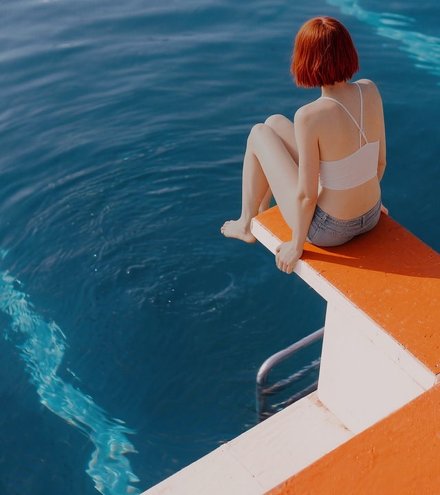 Auf dem Bild ist eine Frau in Bikini zu sehen. Die Frau sitzt die auf einem Schwimmbad-Startblock. Unter ihr ist das dunkelblaue Wasser des Schwimmbeckens zu sehen. Der Startblock ist orange.