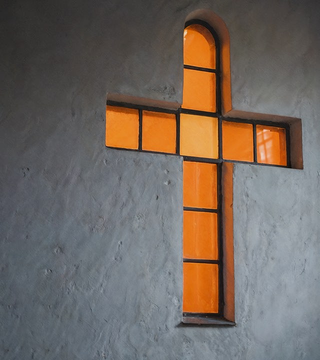 Auf dem Bild ist ein kreuzförmiges oranges Fenster zu sehen. Durch das Fenster scheint Licht nach außen. Das Fenster ist in einer grauen Beton-Wand eingelassen.