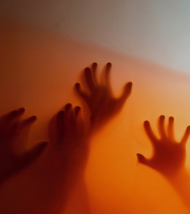 Auf dem Bild ist eine Milchgalsscheibe zu erkennen. Durch die Scheibe sind Schemenhaft vier Hände zu erkennen, die in Richtung der Scheibe greifen. Die Lichtstimmung ist im kompletten Bild orange.