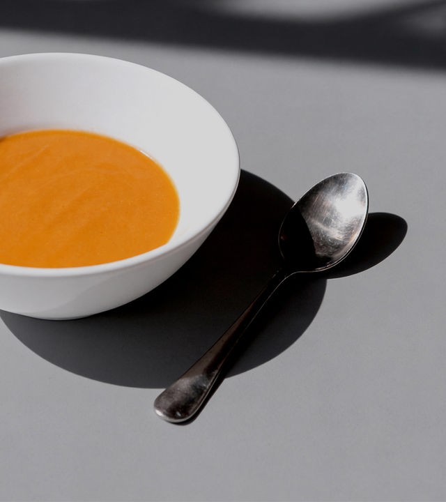 Auf dem Bild ist eine weiße Suppenschüssel zu sehen. In der Suppenschüssel ist orangene Suppe. Rechts neben der Schüssel liegt ein Löffel. Die Schüssel und der Löffel befinden sich auf einem grauen Tisch.