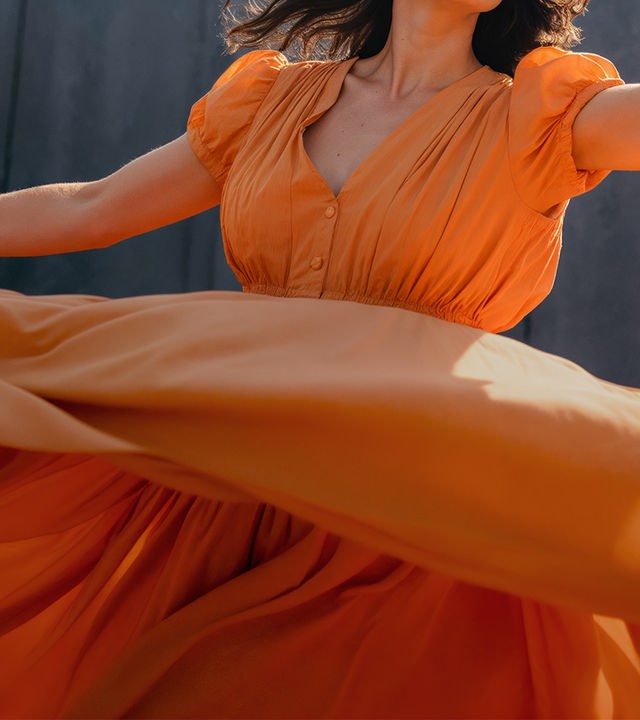 Auf dem Bild ist eine Frau in einem weiten, orangenen Sommerkleid zu sehen. Der Bildausschnitt ist so gewählt, dass das Gesicht ausßerhalb des abgebildeten Bereichs ist. Das Kleid fächert sich breit aus, als würde die Frau sich schnell um ihre eigene Achse drehen.