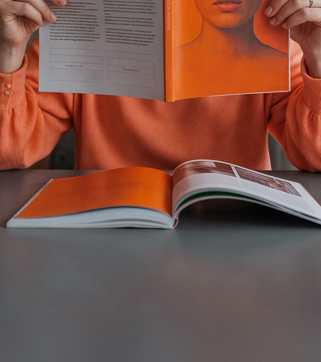 Auf dem Bild ist eine Person im oragenen Pullover zu sehen. Der Bildausschnitt ist so gewählt, dass das Gesicht der Person ausßerhaöb des Bildes ist. Dier Person sitzt an einem Tisch. Sowohl in ihren Händen, als auch vor sich auf dem Tisch befindest sich jeweils ein Magazin. Die überwiegende Farbe in beiden Magazinen ist orange.