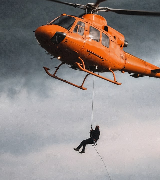 Auf dem Bild ist ein orangener Helikopter abgebildet. Im Hintergrund ist der graue, wolkenverhangene Himmel zu erkennen. Am Helikopter ist ein Seil befästigt, an dem ein Mensch sich abseilt.