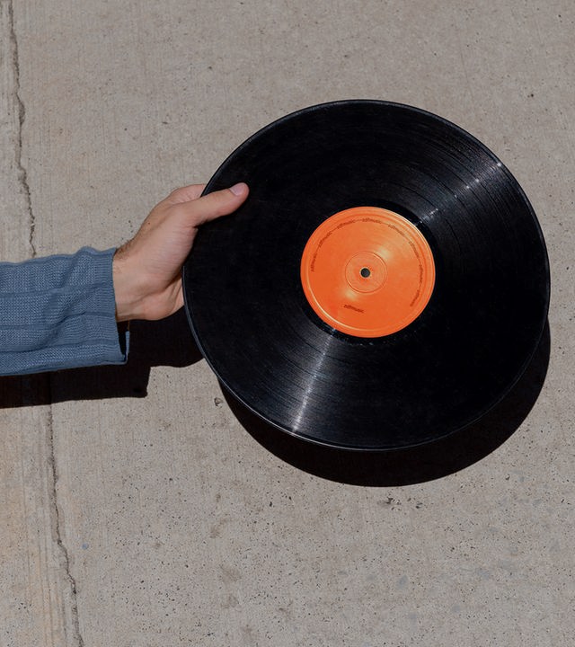 Auf dem Bild ist eine Hand zu erkennen, die eine Schallplatte hält. Das Schallplattenetikett ist orange. Im Hintergrund ist eine graue Betonwand zu erkennen.