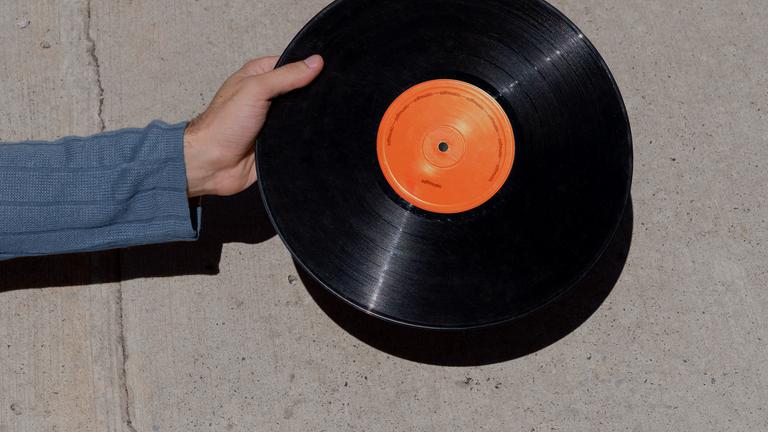 Auf dem Bild ist eine Hand zu erkennen, die eine Schallplatte hält. Das Schallplattenetikett ist orange. Im Hintergrund ist eine graue Betonwand zu erkennen.