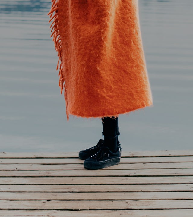 Auf dem Bild ist der Unterkörper einer Person zu sehen. Die Person steht auf einem Steg, derauf eine Wasseroberfläche herausragt. Die Person trägt schwarze Schuhe, eine schwarze Hose und einen orangenen, zotteligen, langen Mantel.
