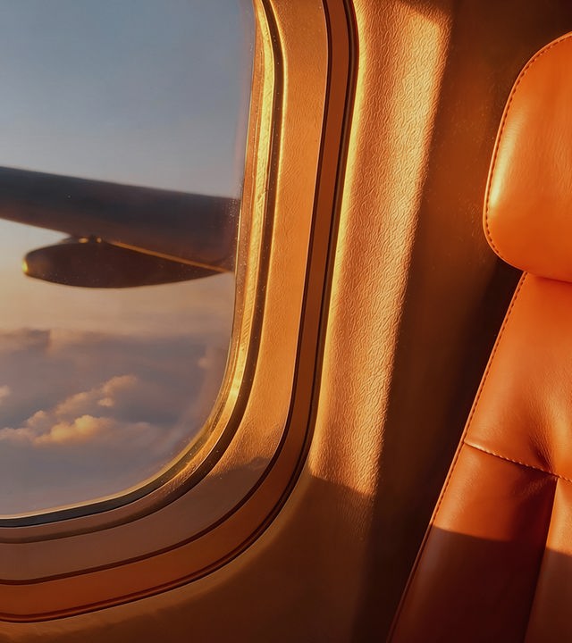 Zu sehen ist ein Sitz in einem Flugzeug. Links vom Sitz ist ein Fenster zu sehen, das den Blick aus dem Flugzeug heraus auf den Flügel und die Wolken gestattet.