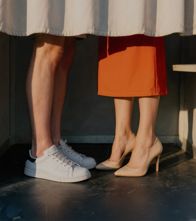 Auf dem Bild sind zwei paar Füße zu sehen. Die Personen zu denen die Füße gehören stehen in einer Passfotokabiene mit zugezogenem Vorhang. Sie stehen einander zugewandt. die linke Person trägt weiße Sneaker und beige Shorts. Die rechte Person trägt beige Schuhe mit hohem Absatz und einen orangenen Midirock.
