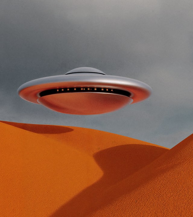 Auf dem Bild ist ein UFO abgebildet. Das UFO schimmert metalisch und spiegelt die Umgebung. Es schwebt über einer orangenen Hügellandschaft. Der Himmer ist grau und wolkenbedeckt.