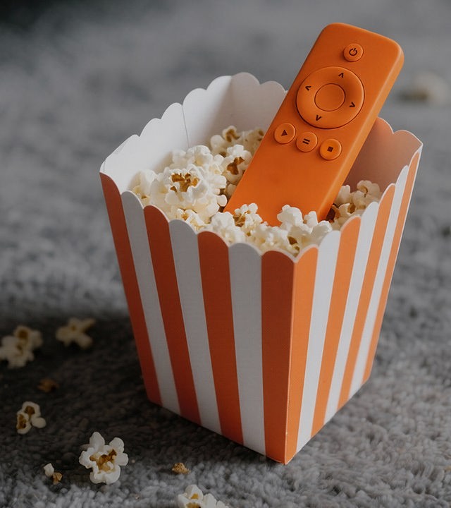 Auf dem Bild ist eine Popcorn-Tüte abgebildet. Die Tüte ist längs orange / weiß gestreift. In der Popcorn-Tüte befindet sich Popcorn und eine orangene Fernbedienung. Die Popcorntüte steht auf einem grauen Teppichboden.