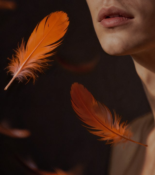 Nahaufnahme des unteren Gesichts und Halses einer Person vor dunklem Hintergrund; mehrere orangefarbene Federn schweben im Vordergrund und erzeugen eine ruhige, sinnliche Stimmung.