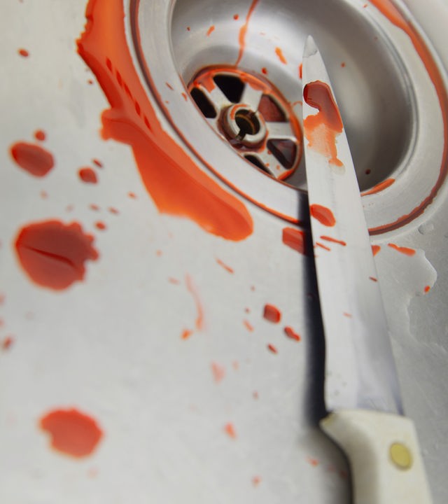 Nahaufnahme eines metalenen Waschbeckens mit Abfluss. Ein Küchenmesser liegt quer über dem Beckenrand, auf Klinge und Oberfläche sind orange Flüssigkeitsspritzer zu sehen die an Blut erinnern und in den Abfluss laufen. Die Szene wirkt kühl und angespannt, der Hintergrund ist unscharf.