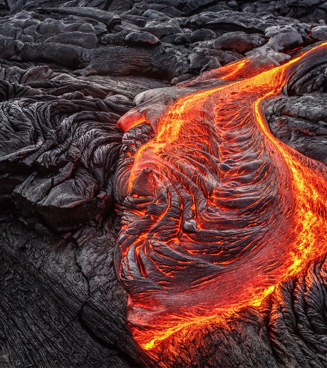 Das Bild zeigt einen Strom aus flüssiger Lava. Die Lava leuchtet orange. Um den Strom flüßige Lava ist viel schon erkaltetes, dunkelgraues Gestein.