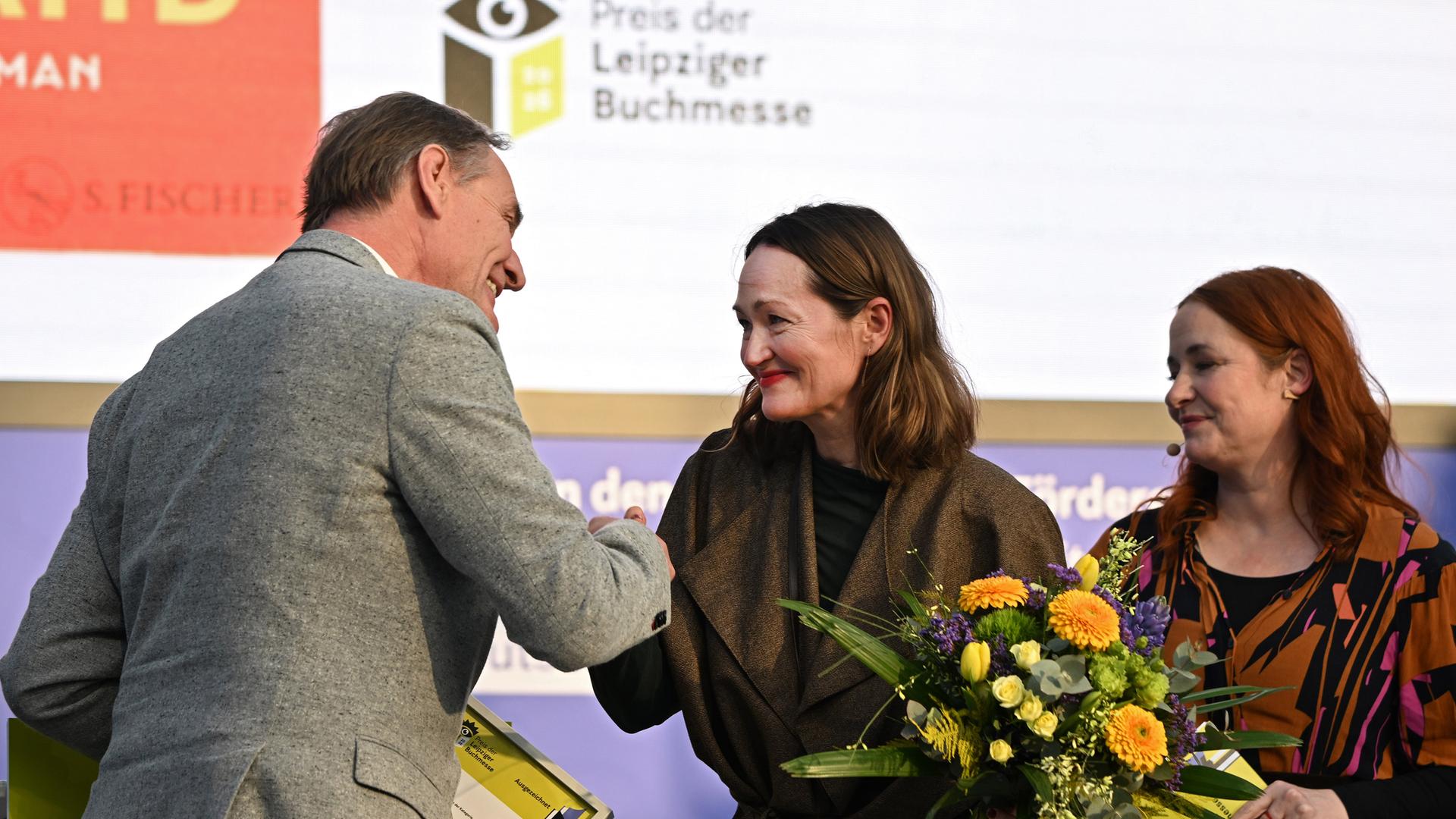 Eine Frau bekommt auf einer Bühne von einem Mann die Hand geschüttelt, neben ihr steht eine Frau mit Blumenstrauß.