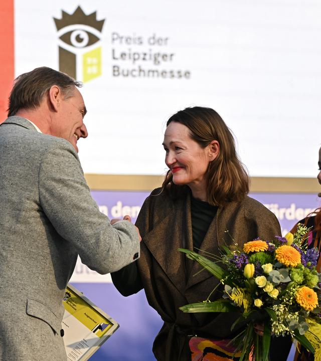 Eine Frau bekommt auf einer Bühne von einem Mann die Hand geschüttelt, neben ihr steht eine Frau mit Blumenstrauß.