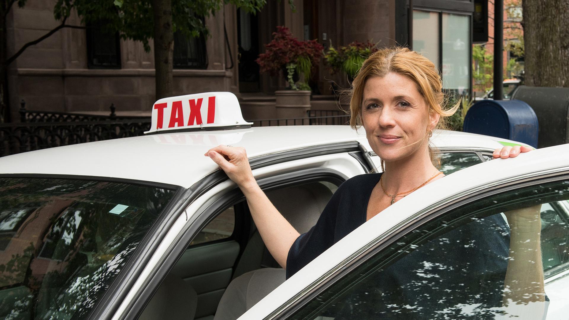 Die Taxifahrerin Audrey (Fiona Coors) steht lächelnd an ihrem weißen Taxi.