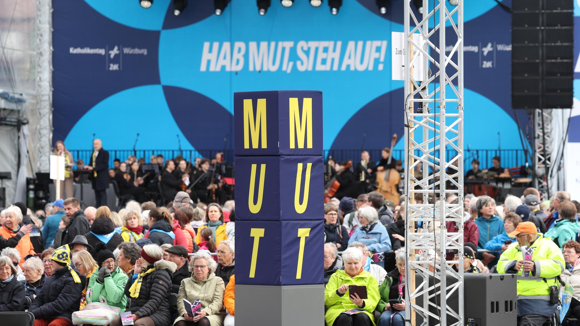 Menschenmasse auf dem Platz, eine Stehle mit der Aufschrift Mut in der Mitte, im Hintergrund das Motto des Katholikentags: Hab Mut, szteh auf!