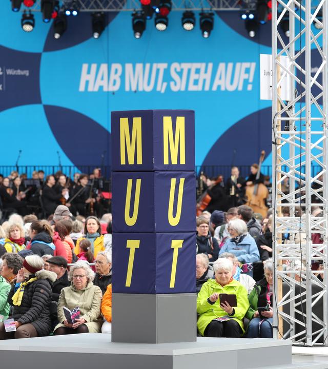 Menschenmasse auf dem Platz, eine Stehle mit der Aufschrift Mut in der Mitte, im Hintergrund das Motto des Katholikentags: Hab Mut, szteh auf!