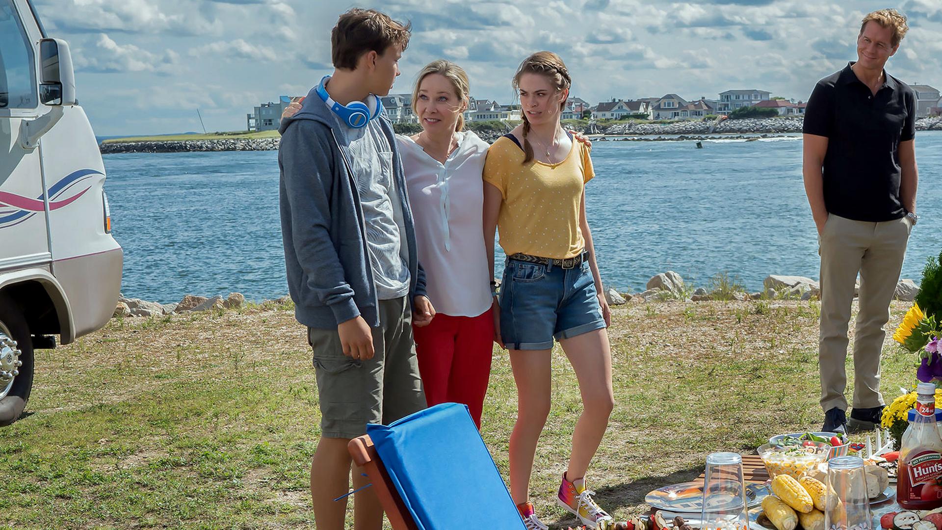 "Katie Fforde: Familie auf Bewährung": Jane Grant (Ann-Kathrin Kramer), ihr Mann Clark (Thomas Heinze) und ihre Kinder Lilly (Pauline Rénevier) und Robin (Sam Heinze) befinden sich auf einem Campingplatz an einem See. Jane steht lachend zwischen ihren Kindern und hat ihre Arme um sie gelegt. Clark steht etwas abseits am Grill und beobachtet seine Familie. Die Sonne scheint, und der Tisch ist gedeckt.