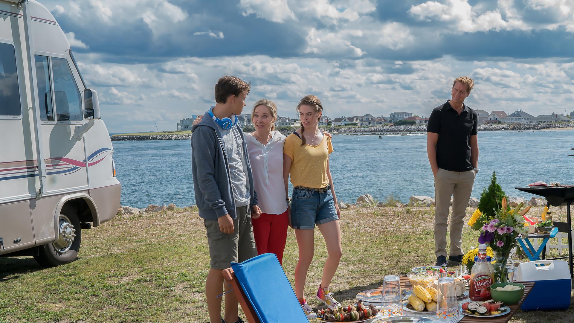 "Katie Fforde: Familie auf Bewährung": Jane Grant (Ann-Kathrin Kramer), ihr Mann Clark (Thomas Heinze) und ihre Kinder Lilly (Pauline Rénevier) und Robin (Sam Heinze) befinden sich auf einem Campingplatz an einem See. Jane steht lachend zwischen ihren Kindern und hat ihre Arme um sie gelegt. Clark steht etwas abseits am Grill und beobachtet seine Familie. Die Sonne scheint, und der Tisch ist gedeckt.