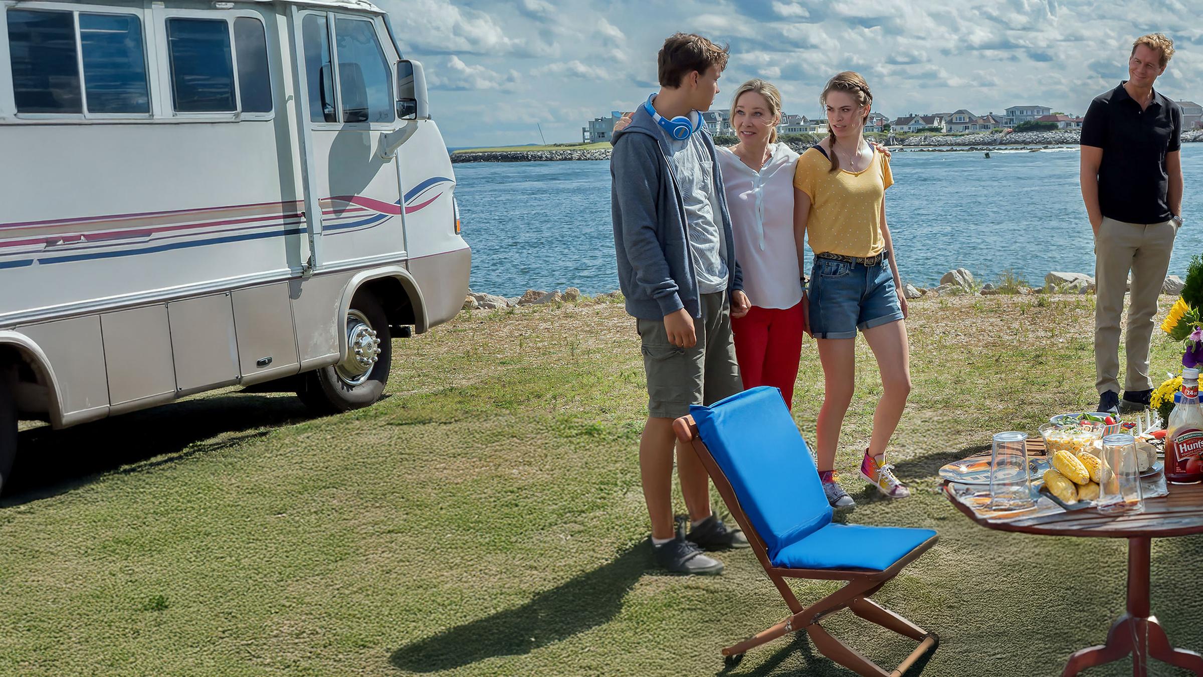 "Katie Fforde: Familie auf Bewährung": Jane Grant (Ann-Kathrin Kramer), ihr Mann Clark (Thomas Heinze) und ihre Kinder Lilly (Pauline Rénevier) und Robin (Sam Heinze) befinden sich auf einem Campingplatz an einem See. Jane steht lachend zwischen ihren Kindern und hat ihre Arme um sie gelegt. Clark steht etwas abseits am Grill und beobachtet seine Familie. Die Sonne scheint, und der Tisch ist gedeckt.