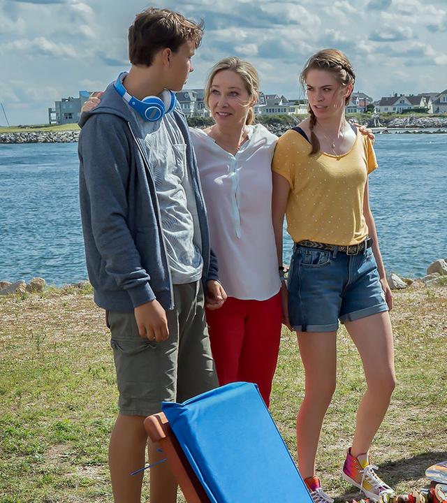 "Katie Fforde: Familie auf Bewährung": Jane Grant (Ann-Kathrin Kramer), ihr Mann Clark (Thomas Heinze) und ihre Kinder Lilly (Pauline Rénevier) und Robin (Sam Heinze) befinden sich auf einem Campingplatz an einem See. Jane steht lachend zwischen ihren Kindern und hat ihre Arme um sie gelegt. Clark steht etwas abseits am Grill und beobachtet seine Familie. Die Sonne scheint, und der Tisch ist gedeckt.