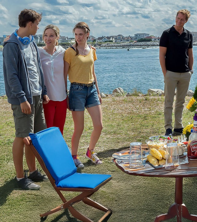 "Katie Fforde: Familie auf Bewährung": Jane Grant (Ann-Kathrin Kramer), ihr Mann Clark (Thomas Heinze) und ihre Kinder Lilly (Pauline Rénevier) und Robin (Sam Heinze) befinden sich auf einem Campingplatz an einem See. Jane steht lachend zwischen ihren Kindern und hat ihre Arme um sie gelegt. Clark steht etwas abseits am Grill und beobachtet seine Familie. Die Sonne scheint, und der Tisch ist gedeckt.