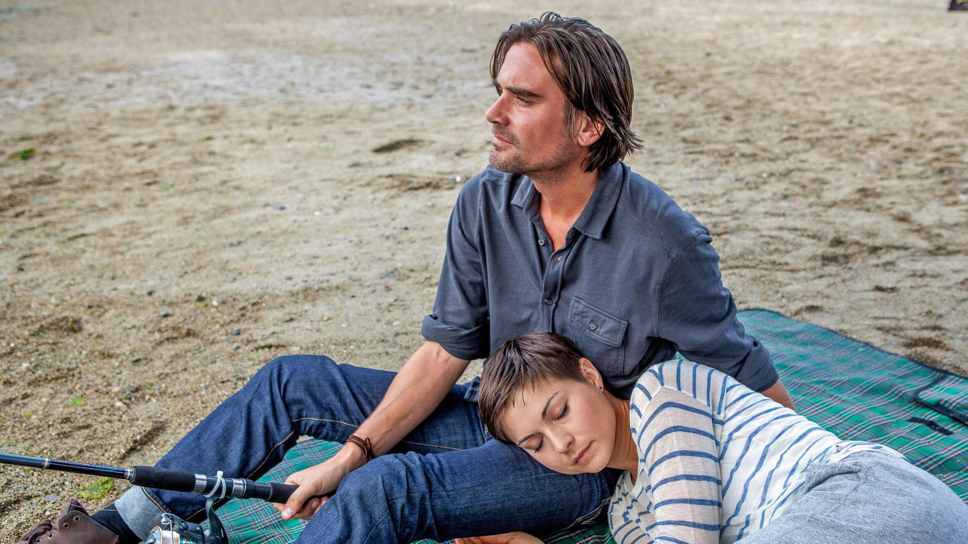 "Katie Fforde: Vergissmeinnicht": Natalie Wayne (Henriette Richter-Röhl) und Matthew Bradford (Patrick Rapold) befinden sich am Strand. Sie liegt mit ihrem Kopf, die Augen geschlossen, auf seinem Bein. Er hält eine Angel in der Hand und schaut in die Ferne.