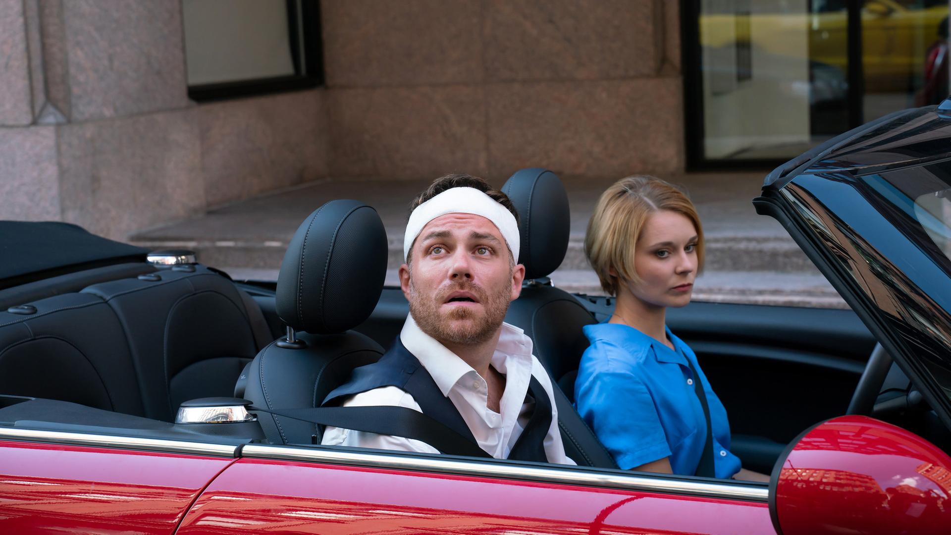 "Katie Fforde: Wachgeküsst": Maggie (Nadja Bobyleva) sitzt im Auto besorgt neben Chris (Bernhard Piesk), der einen Kopfverband trägt und ratlos auf ein Gebäude schaut.