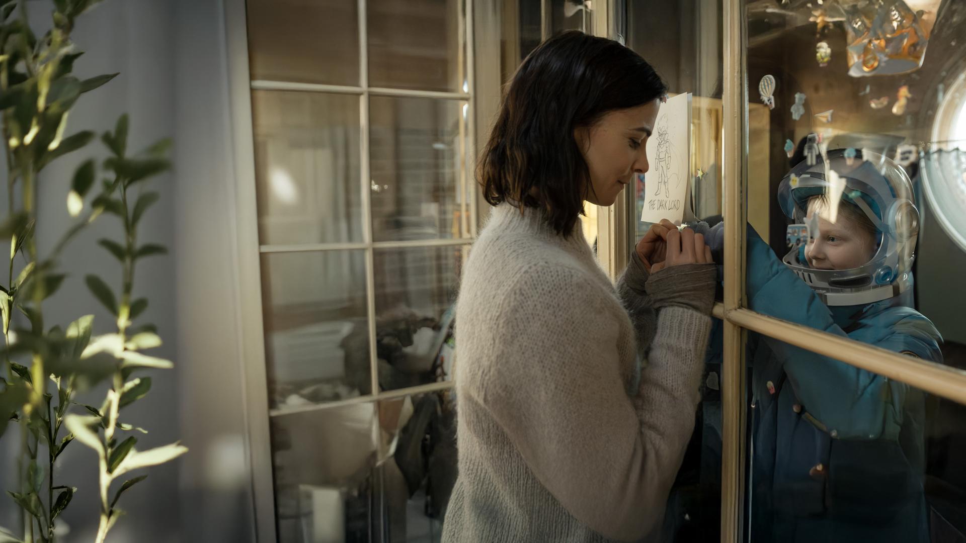 "Serial Cleaner - Kein Zurück": Teri Marquard (Aylin Tezel) und ihre schwerkranke Tochter Luca (Hanna Heckt) befinden sich in einem Raum und sind durch eine Glasscheibe getrennt. Teri hat ihre Hände zu einem Herz geformt und hält sie für ihre Tochter an die Glasscheibe. Luca trägt Schutzkleidung und schaut lächelnd zu ihrer Mutter.