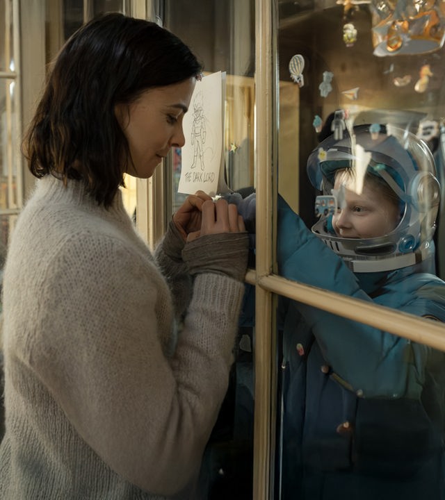 "Serial Cleaner - Kein Zurück": Teri Marquard (Aylin Tezel) und ihre schwerkranke Tochter Luca (Hanna Heckt) befinden sich in einem Raum und sind durch eine Glasscheibe getrennt. Teri hat ihre Hände zu einem Herz geformt und hält sie für ihre Tochter an die Glasscheibe. Luca trägt Schutzkleidung und schaut lächelnd zu ihrer Mutter.