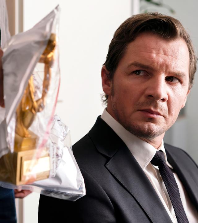 Ein Mann sitzt auf einem Stuhl und betrachtet eine in eine Plastiktüte verpackte Statue, die von einer Hand gehalten wird.