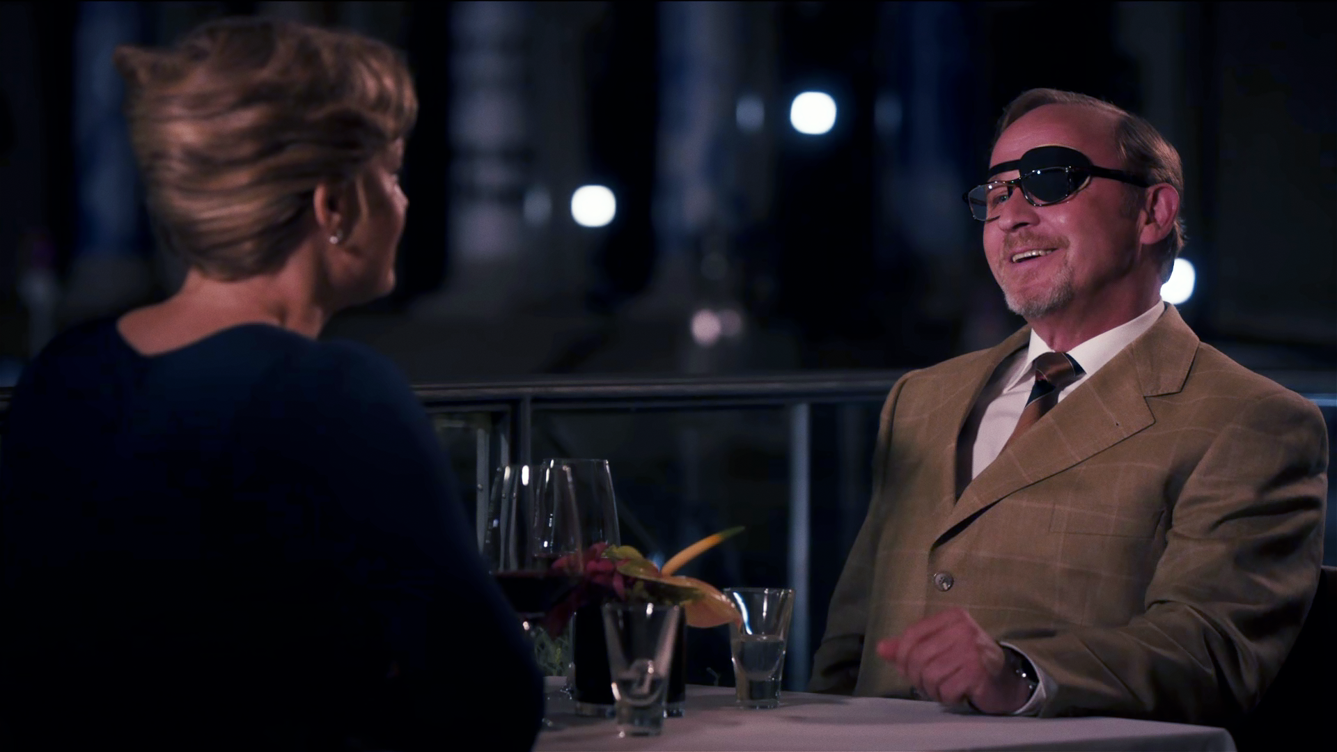 Ludwig Schaller (Alexander Held) sitzt in einem Restaurant einer Frau gegenüber, die nur von hinten zu sehen ist. Er lacht und trägt über dem linken Auge eine Augenklappe. 