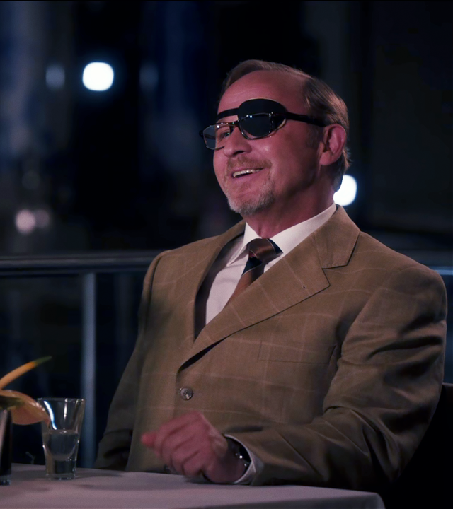 Ludwig Schaller (Alexander Held) sitzt in einem Restaurant einer Frau gegenüber, die nur von hinten zu sehen ist. Er lacht und trägt über dem linken Auge eine Augenklappe. 