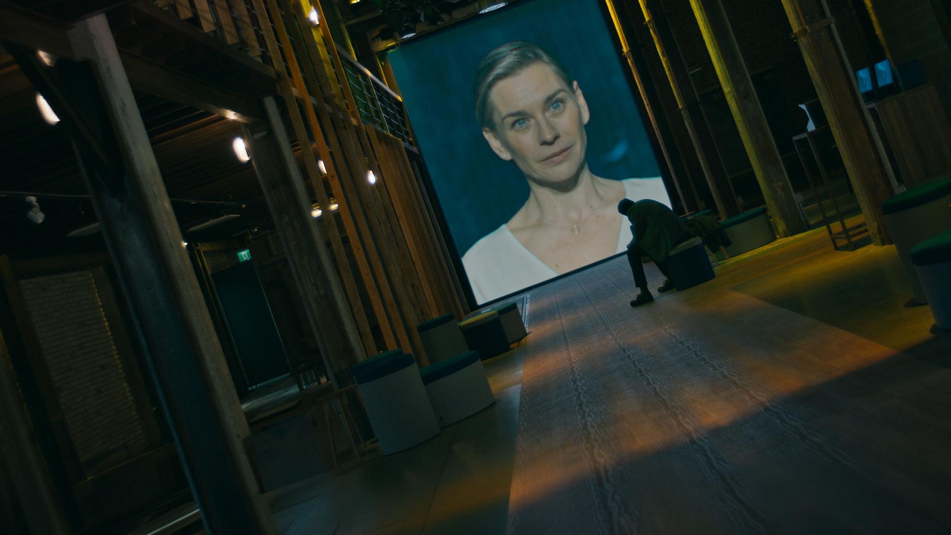 "Concordia - Tödliche Utopie (6) - Kein Weg zurück": Juliane Ericksen (Christiane Paul) ist auf eine Leinwand projiziert. Davor sitzt Noah Ericksen (Steven Sowah) in einem dunklen Raum und sieht zu.